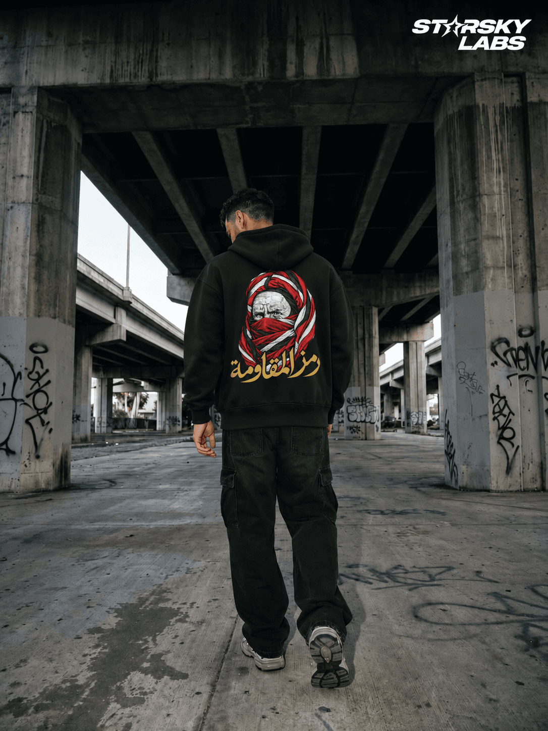 Homme en hoodie noir marchant sous une structure en béton, avec le motif rouge, blanc et doré visible dans le dos.