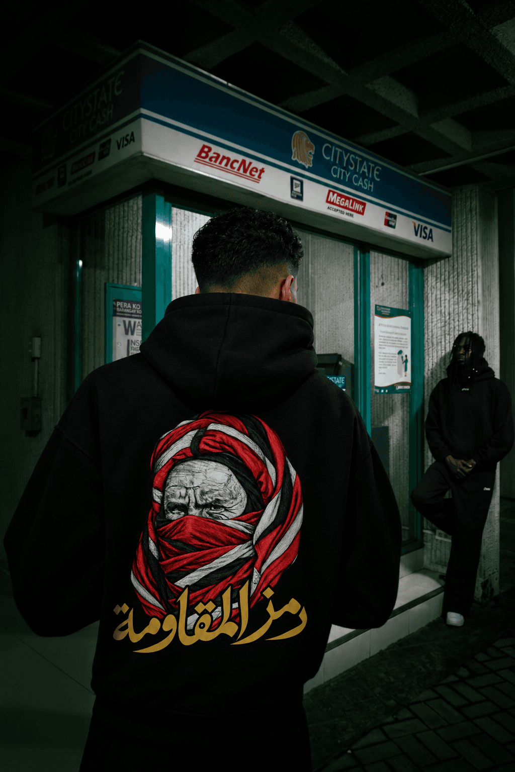 Silhouette de dos devant une façade sombre, portant le hoodie noir au portrait enveloppé de rouge et à la calligraphie dorée.