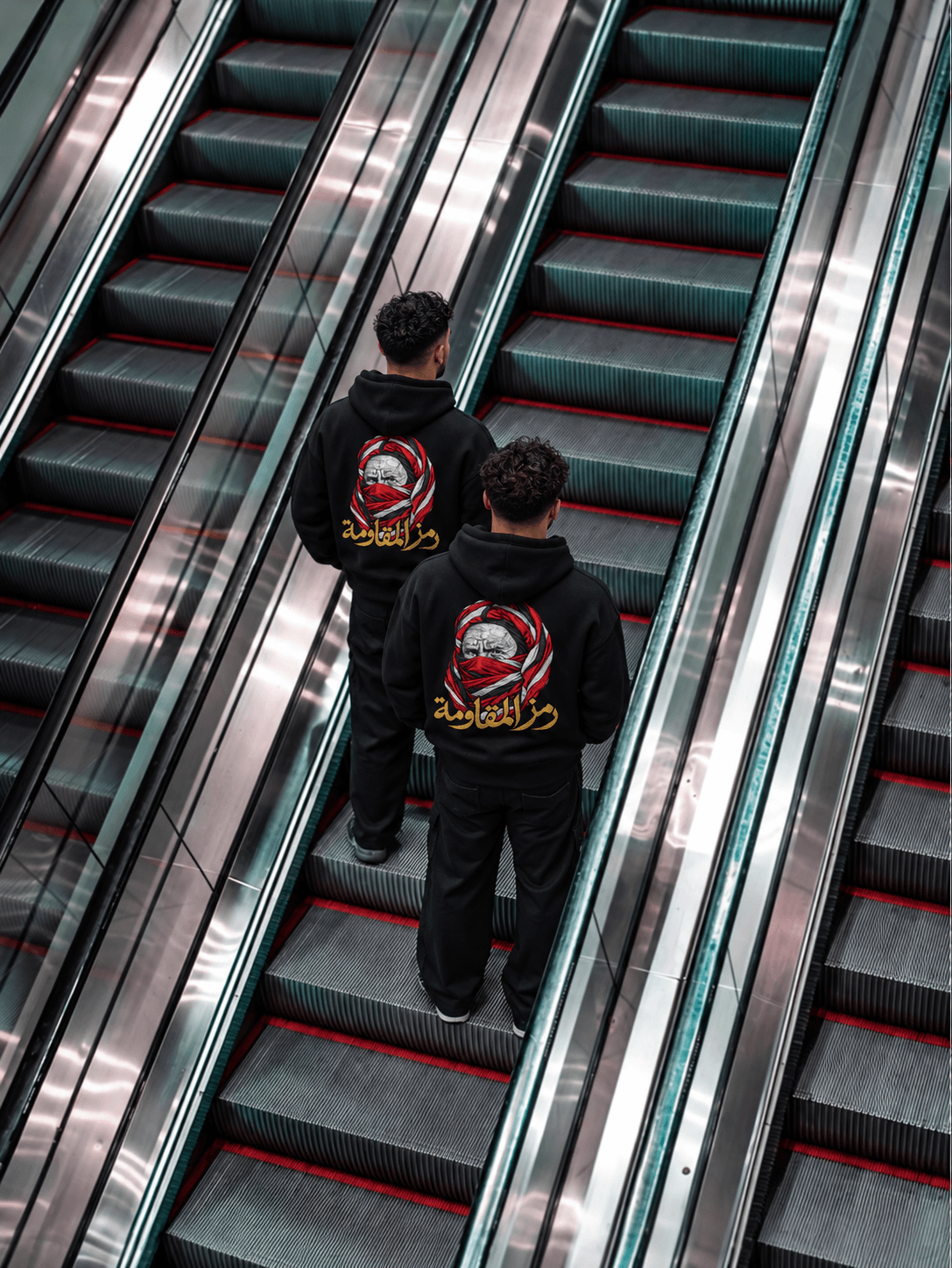 Deux silhouettes de dos dans des escalators, portant le même hoodie noir avec son motif rouge, blanc et doré.