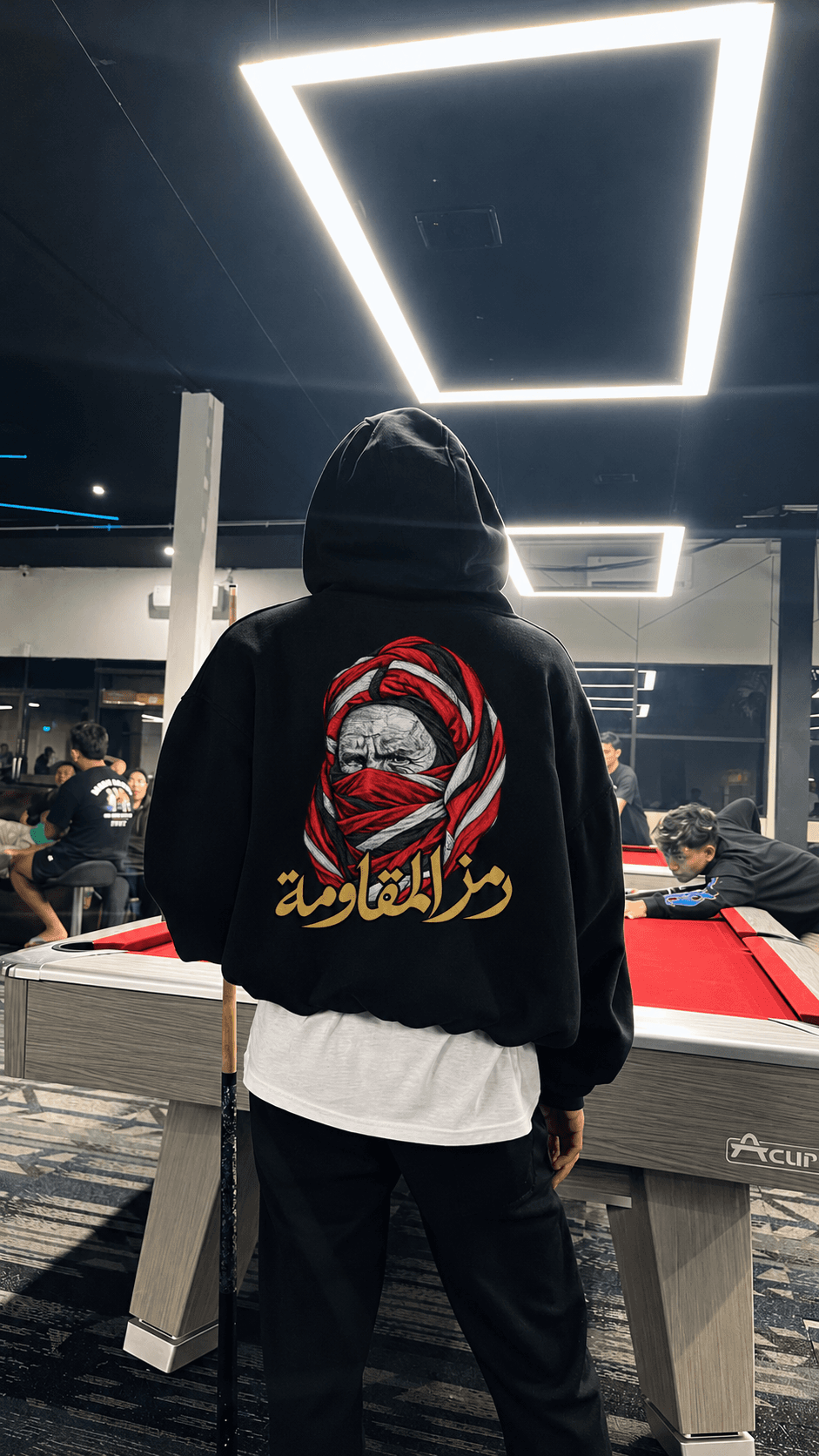 Personne de dos, capuche relevée, dans une salle de billard éclairée par de grands rectangles lumineux, avec le dessin du hoodie visible.