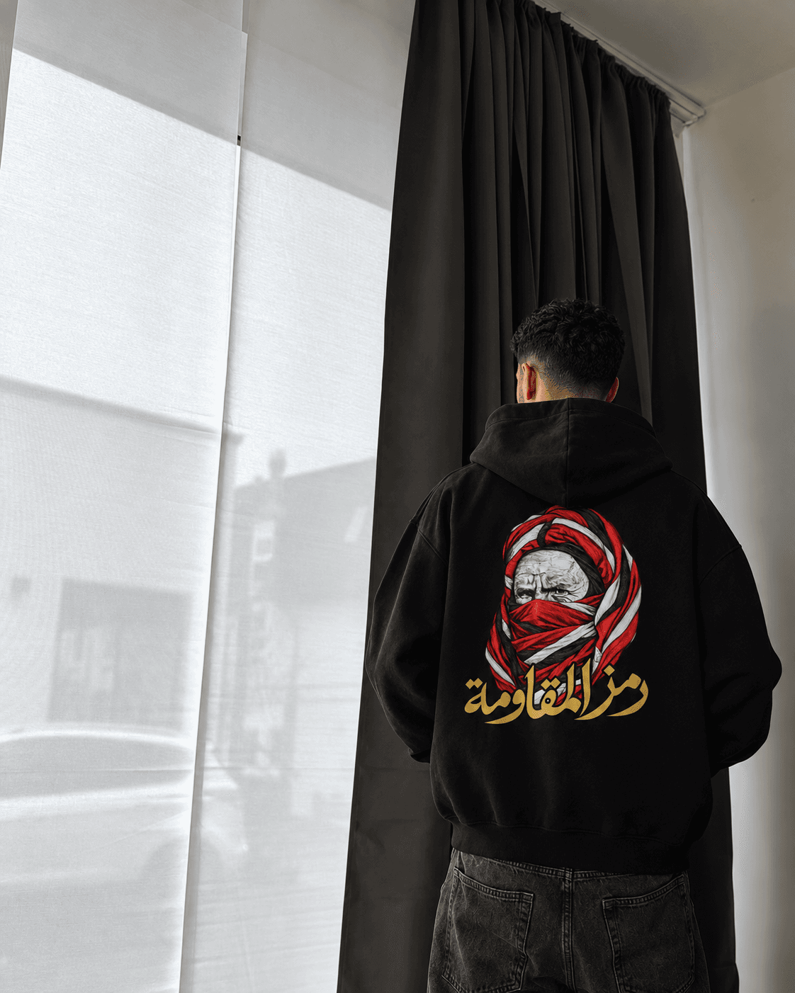 Homme de dos près d’une fenêtre et de rideaux, la lumière naturelle révélant le motif rouge, blanc et doré du hoodie noir.