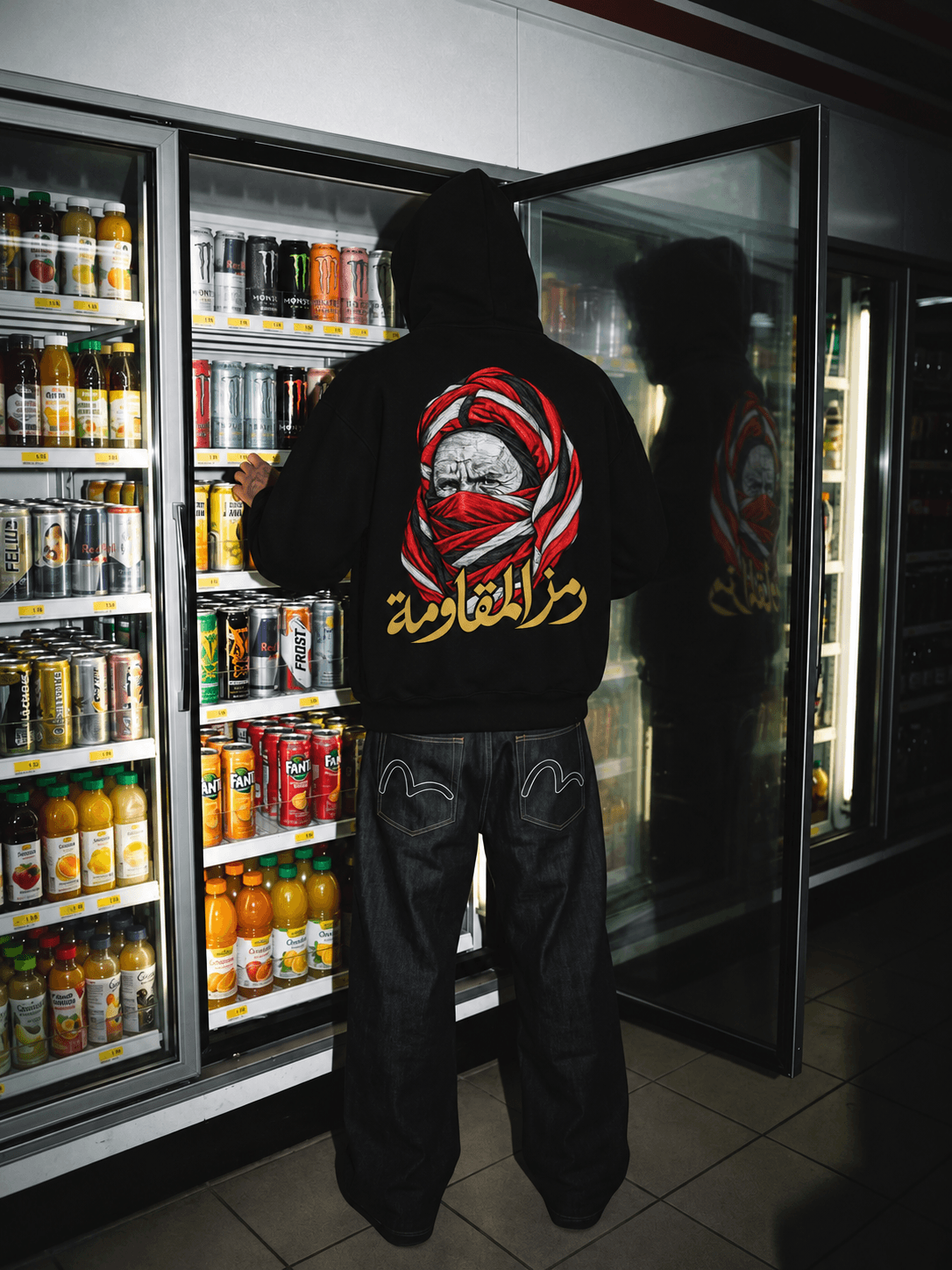 Personne en hoodie noir devant un rayon de boissons, avec le portrait et la calligraphie dorée du dos reflétés dans la porte vitrée ouverte.
