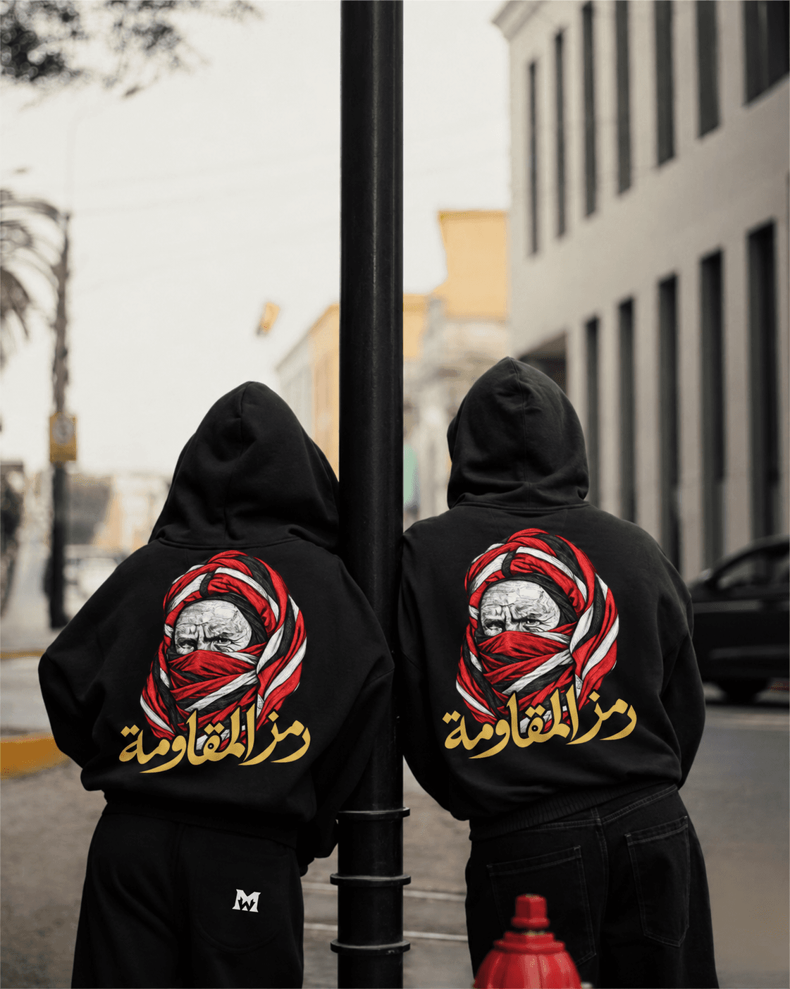 Deux personnes de dos, capuches relevées, de chaque côté d’un lampadaire, portant le même hoodie noir au motif rouge, blanc et doré.