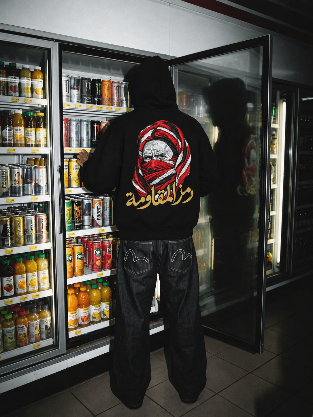 Silhouette en hoodie noir devant des boissons réfrigérées, le portrait rouge et blanc et la calligraphie dorée se reflétant dans une porte vitrée ouverte.