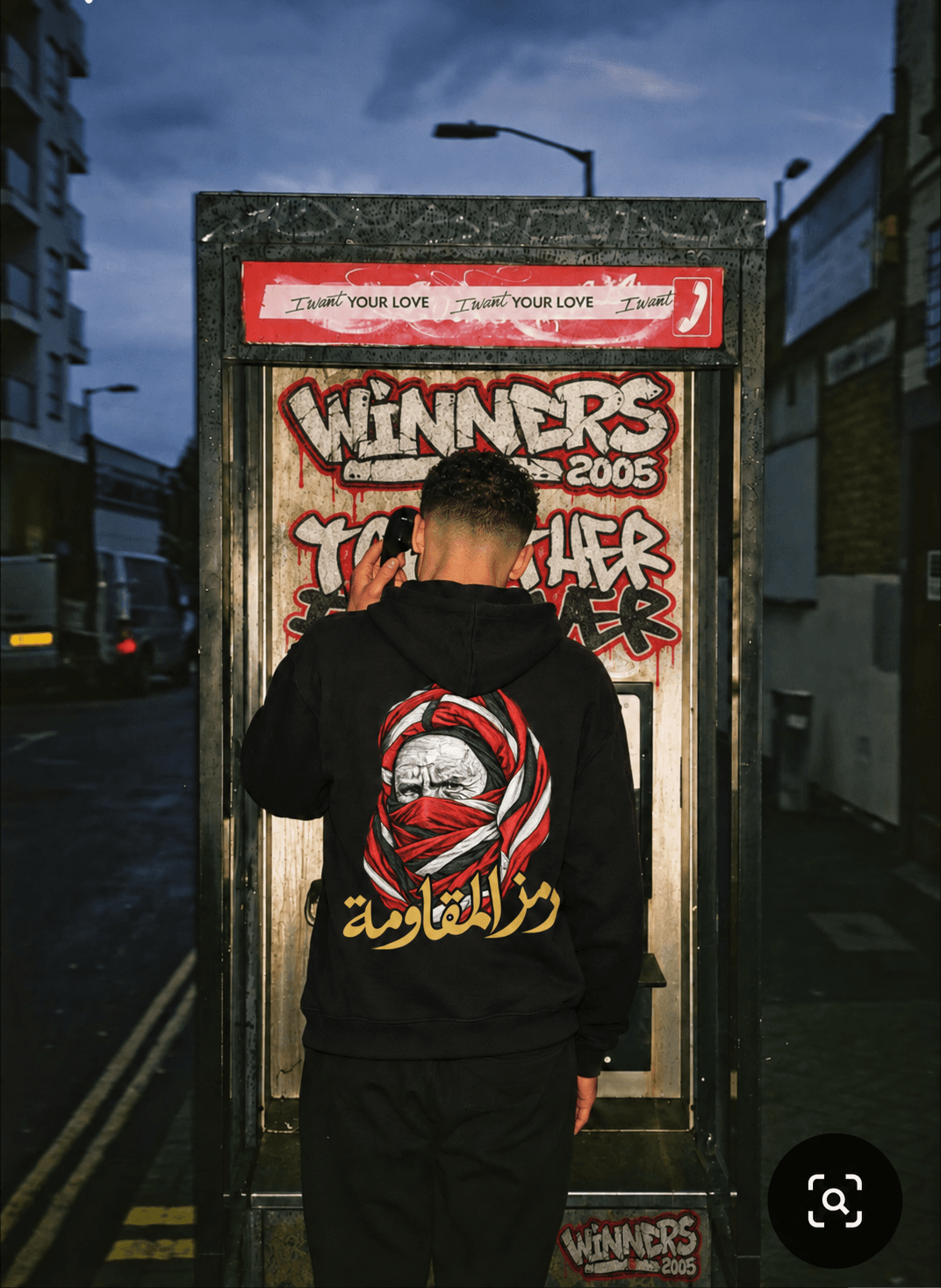 Homme de dos en hoodie noir dans une cabine téléphonique ornée de graffitis WINNERS 2005 et TOGETHER FOREVER, le motif doré visible au dos.