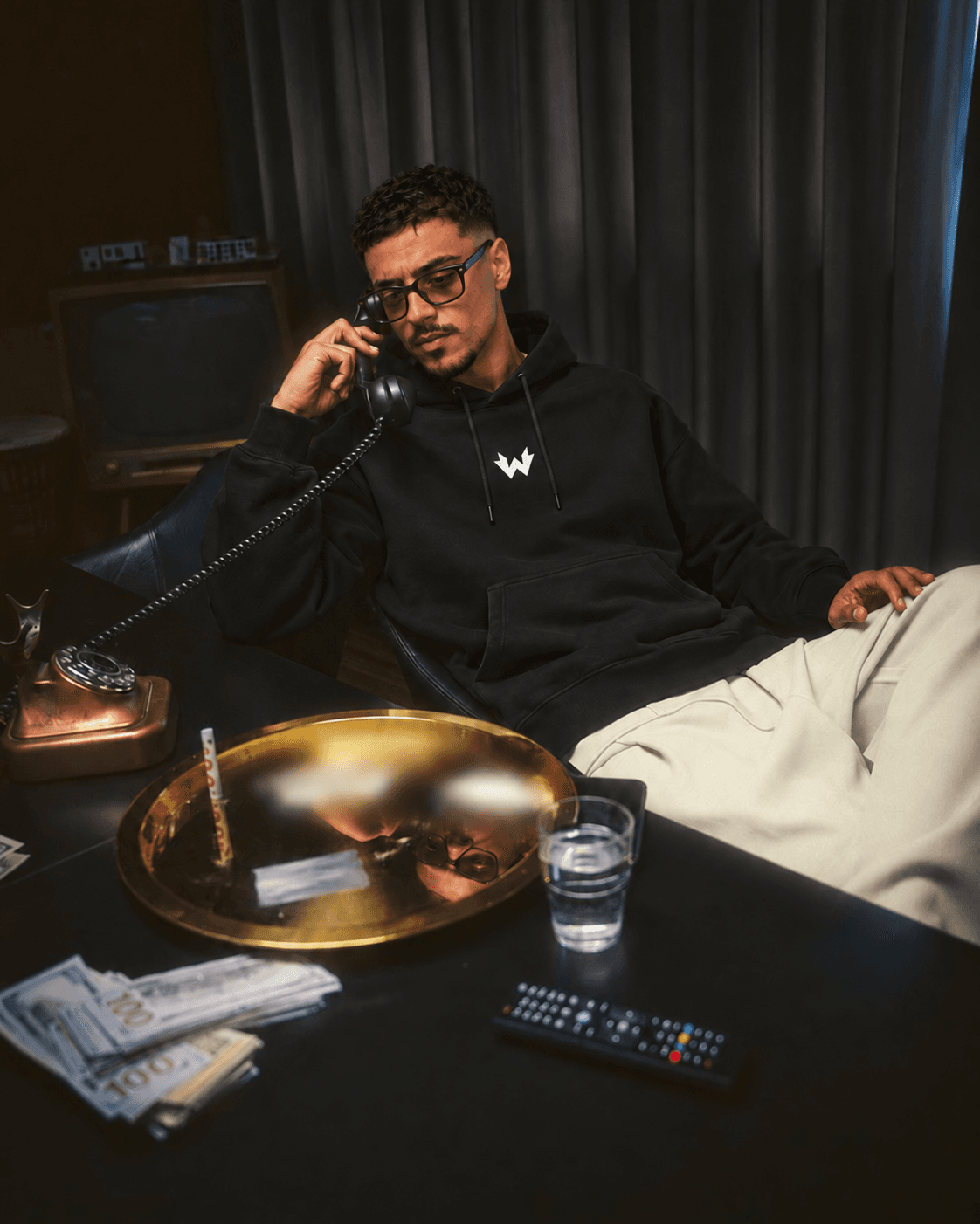Homme à lunettes assis avec un combiné téléphonique, portant le hoodie noir de face avec son petit symbole W blanc sur la poitrine.