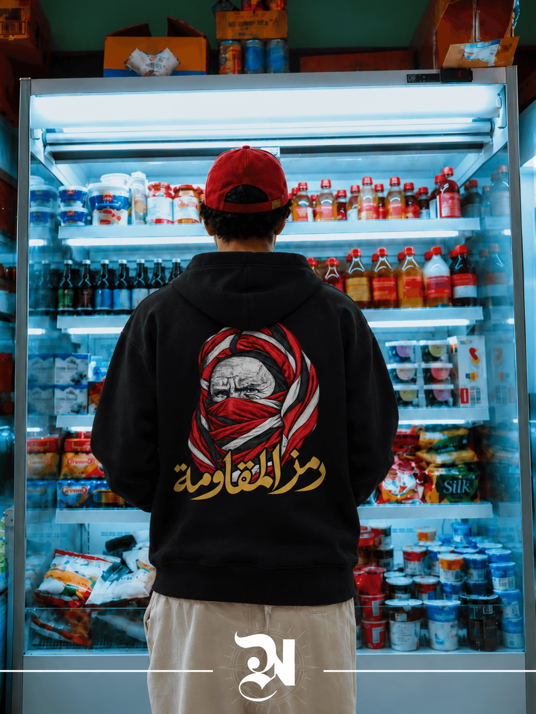 Silhouette de dos en hoodie noir et casquette rouge devant un rayon réfrigéré, avec le portrait rouge et blanc et la calligraphie dorée au dos.